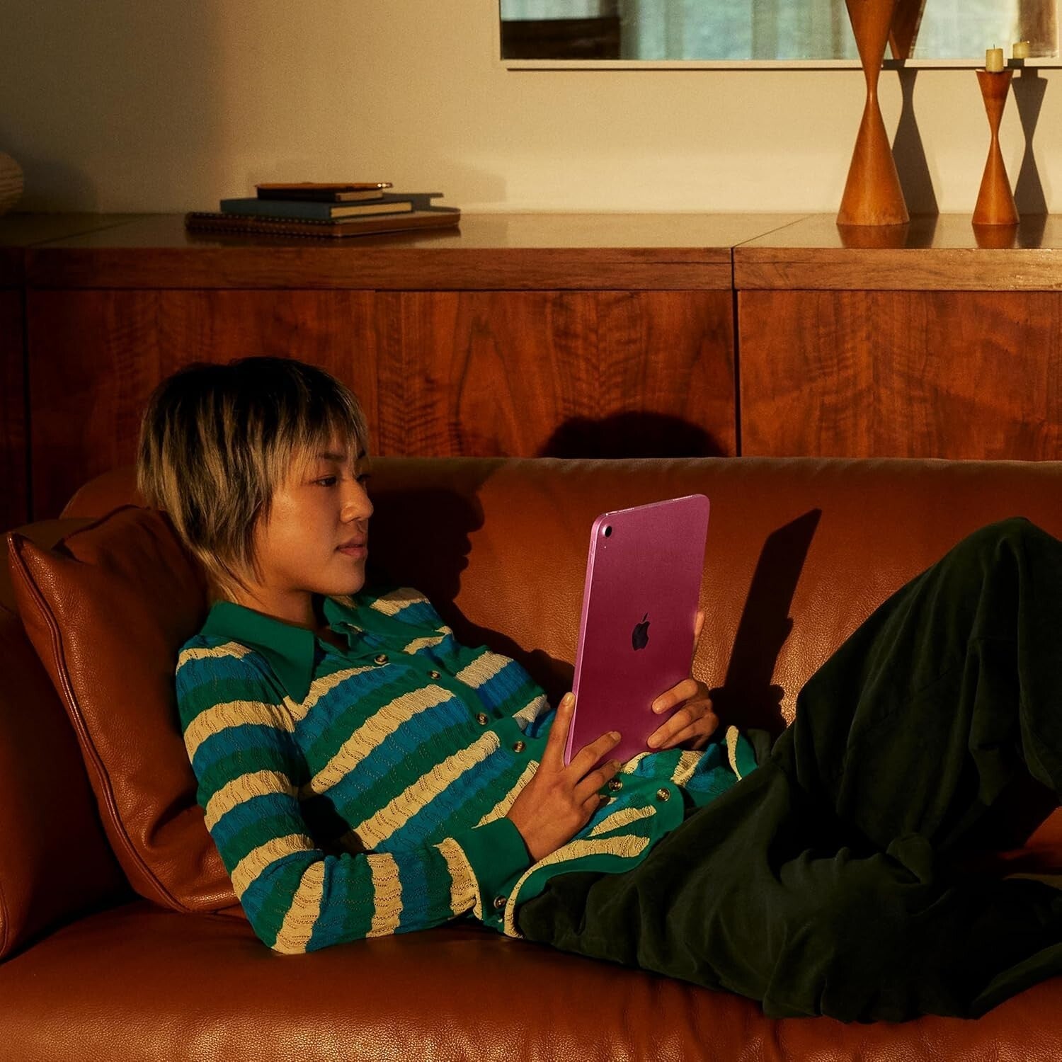 Person reclining on brown leather sofa using pink 11-inch iPad 11th Gen with Apple logo visible in warm indoor lighting
