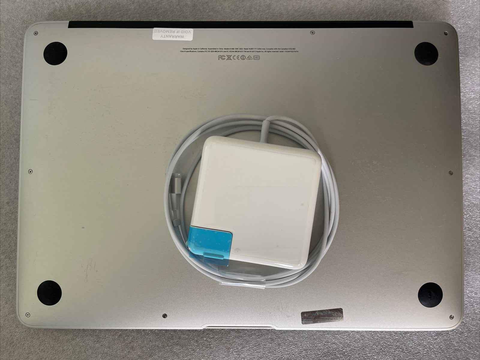 Bottom view of silver MacBook Air 13 inch 2013 model with charger placed on top showing rubber feet and label details