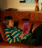Person reclining on brown leather couch using pink Apple iPad 11-inch 11th Gen with visible rear Apple logo in warm lighting
