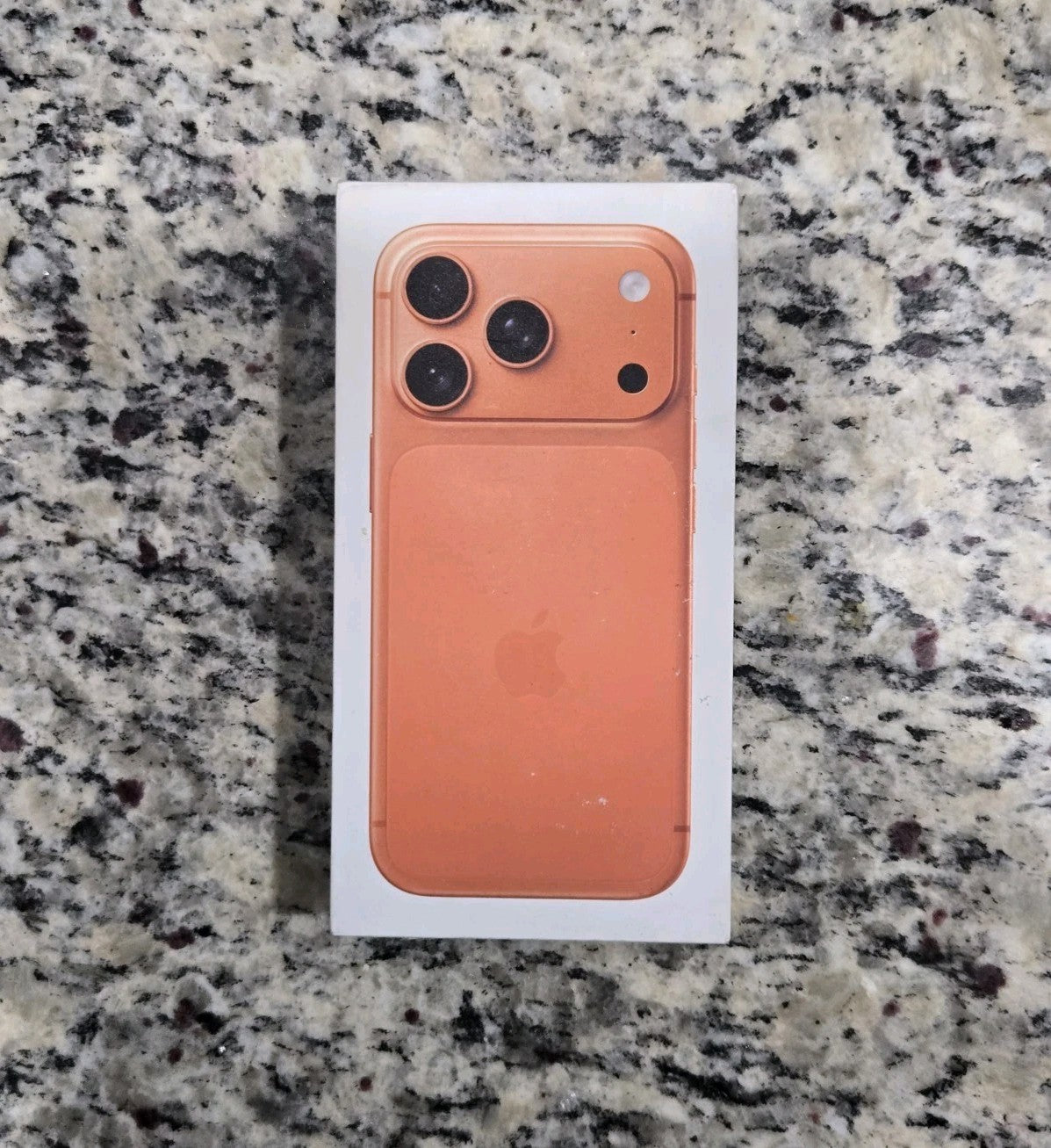 Apple iPhone 17 Pro Cosmic Orange box showing back with triple camera system and Apple logo on granite surface