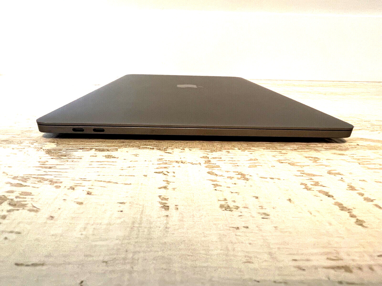 Closed Apple MacBook Pro 15-inch in space gray showing left side with dual Thunderbolt 3 ports and Apple logo on wooden surface