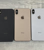 Three Apple iPhone XS Max phones in black, gold, and silver colors laid flat on a wooden surface.