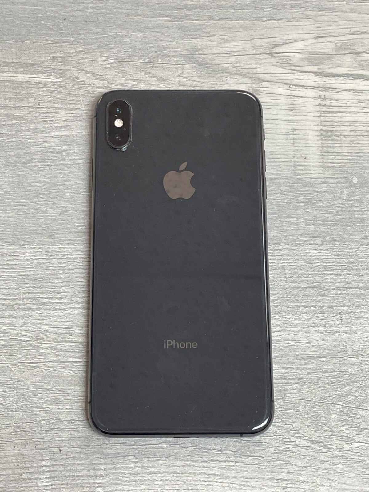 Black Apple iPhone XS Max with dual rear cameras placed on a gray wooden surface showing back view design and logo.